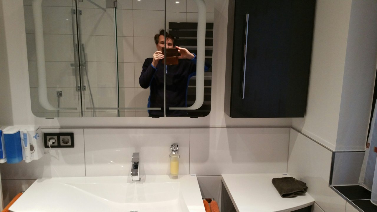 Hängender Spiegelschrank mit abgerundeten Ecken und integrierter Ringbeleuchtung, daneben ein zum Waschtischunterschrank passender Hängeschrank